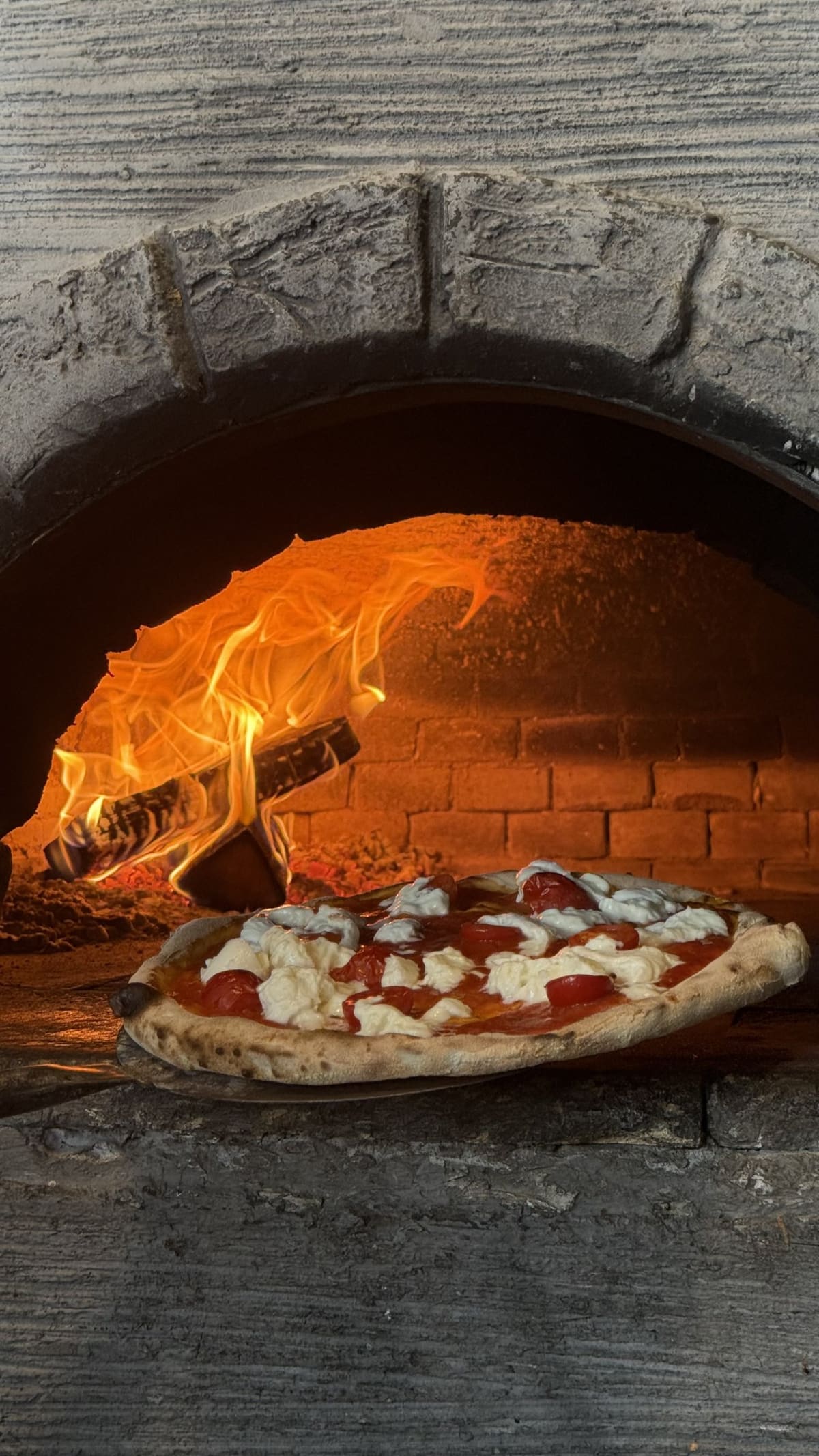 Pizza wird auf dem Schieber in den brennenden Holzofen geschoben